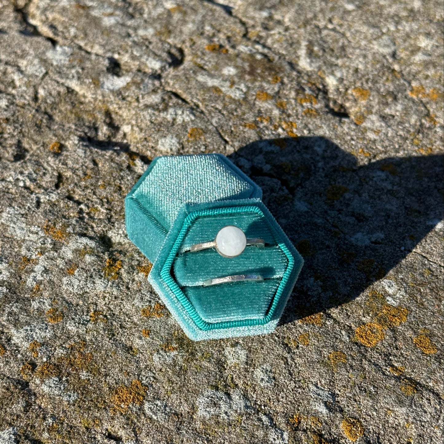 Teal hexagonal jewelry box with a moonstone silverring and a hammered silverring on a textured stone surface