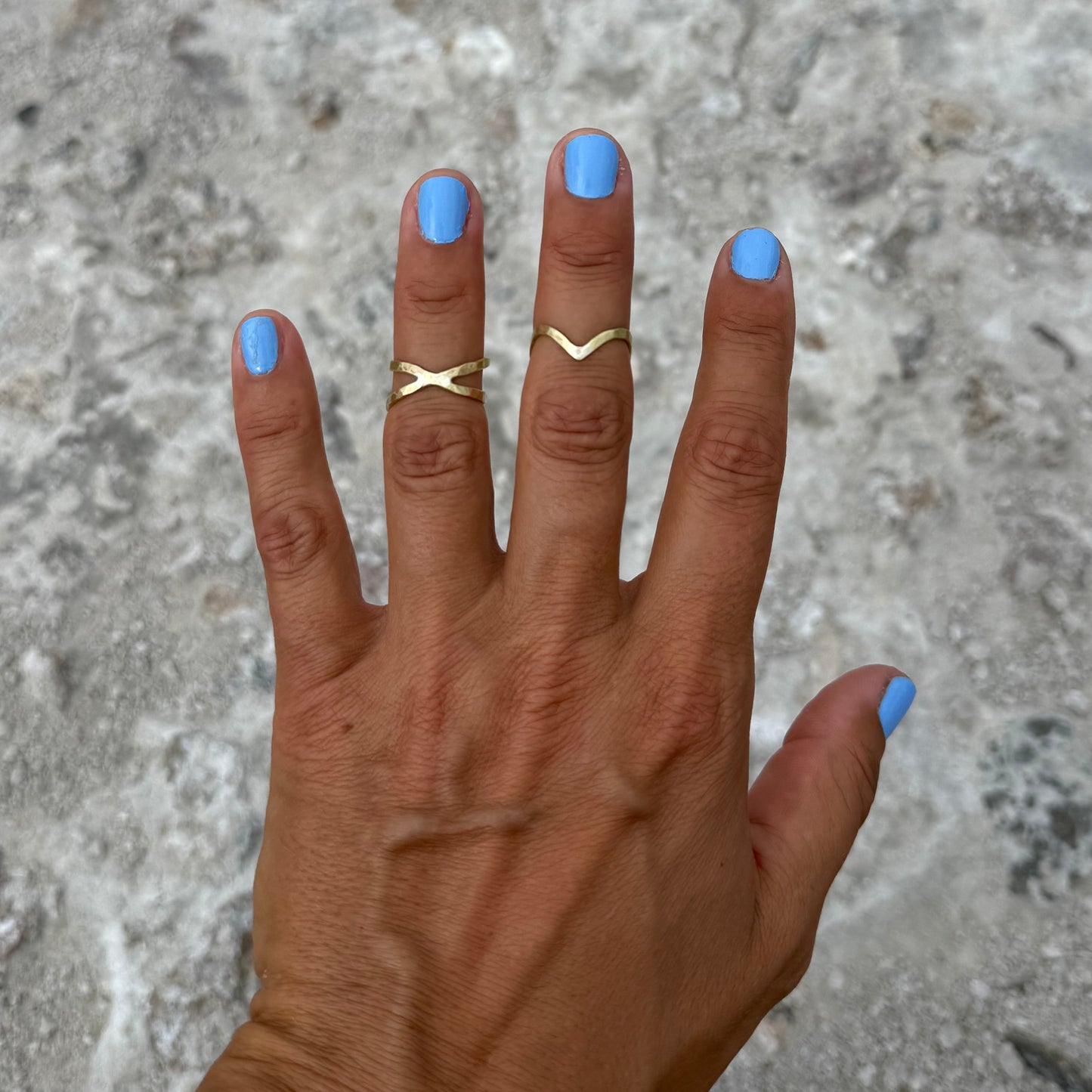 Hand with blue nail polish and a wave-shaped brass ring and infinity brass ring
on a textured stone surface