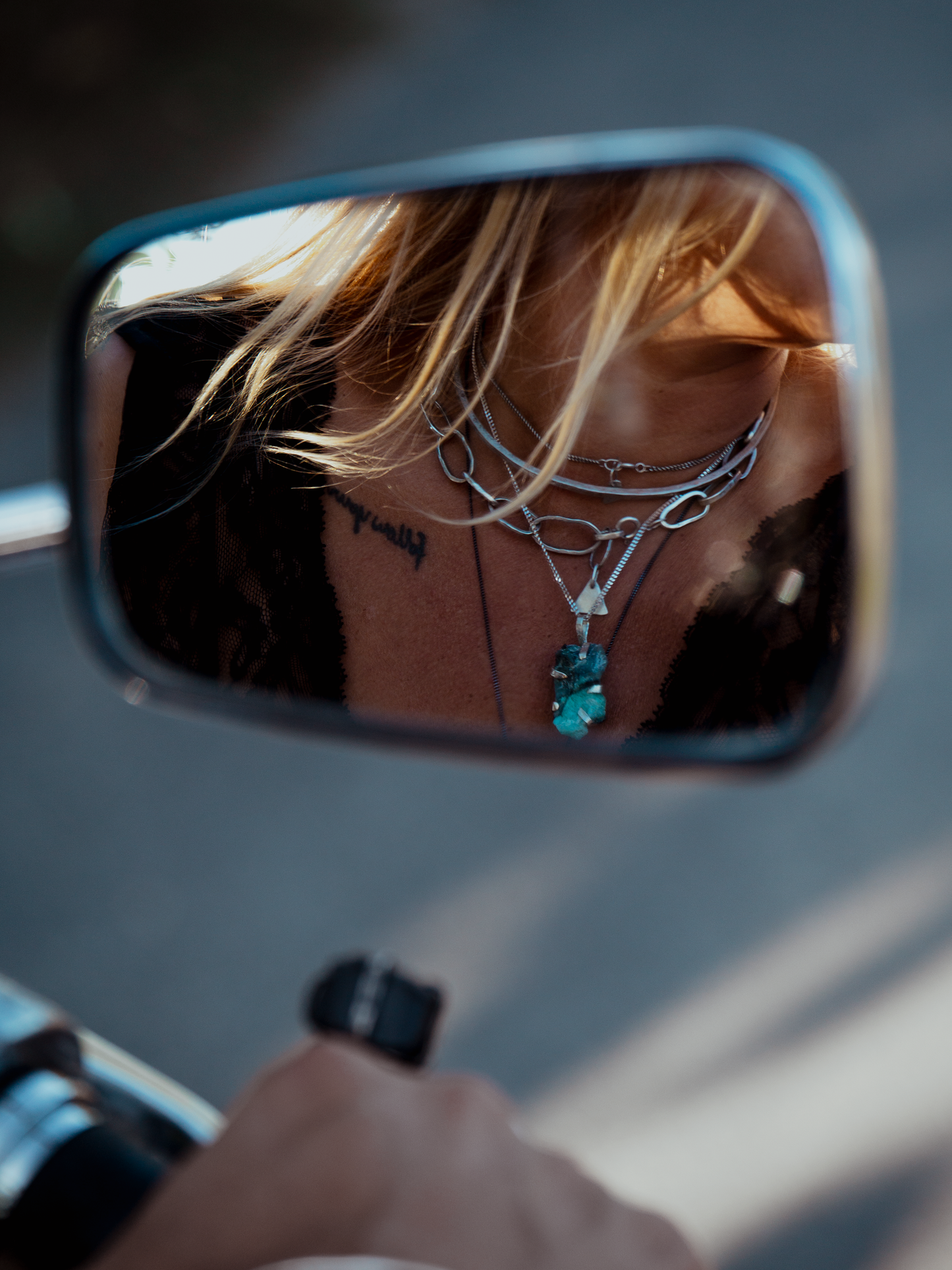 Person wearing necklaces with a blue pendant in a motorcycle's side mirror reflection