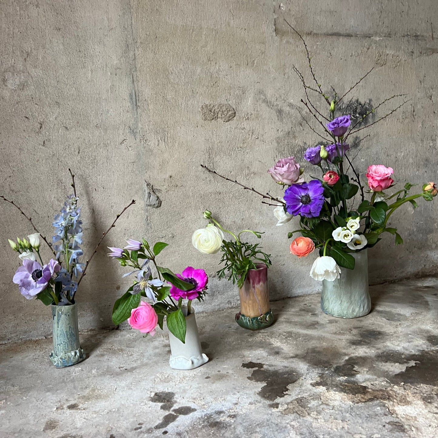 Collection of floral arrangements in various handmade one-of-a-kind ceramics vases on a concrete floor with a textured wall background.