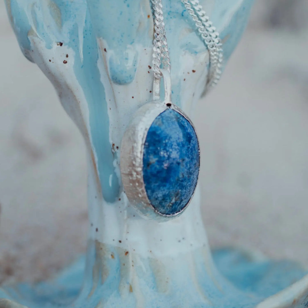 Blue lapis lazuli pendant on a silver chain against a ceramic jewelry stand