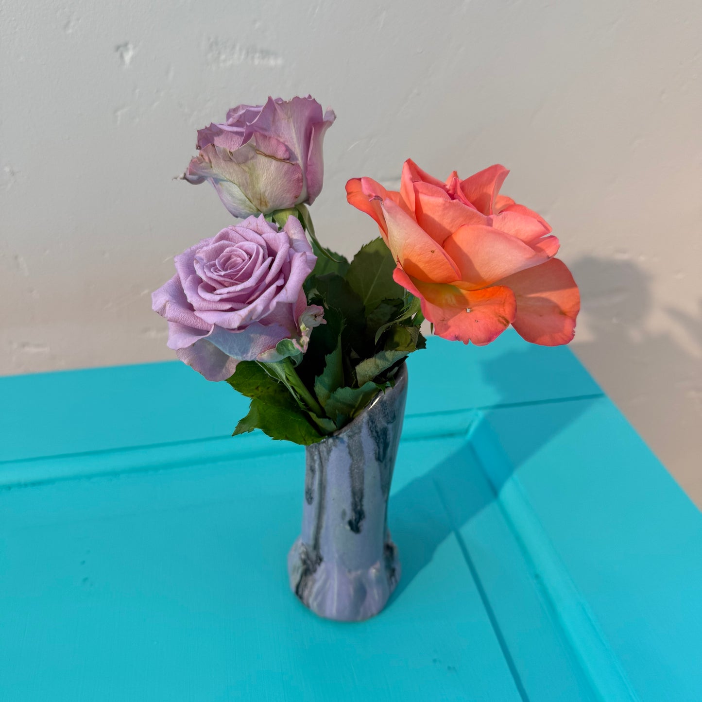 Three artificial roses in a small ceramics vase in purple and black glazing on a blue surface with a white wall background.