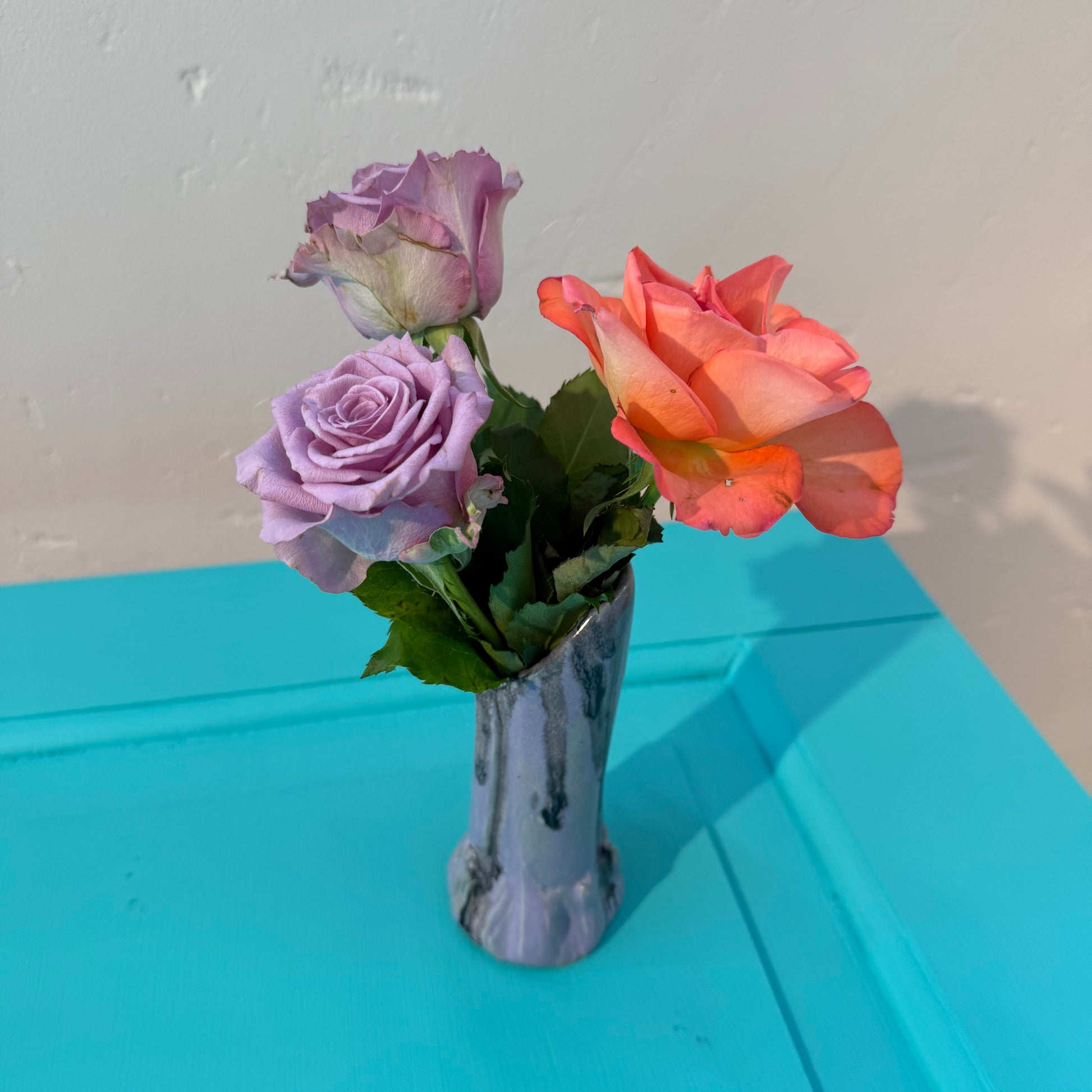 Three artificial roses in a small ceramics vase in purple and black glazing on a blue surface with a white wall background.
