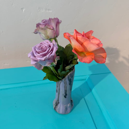 Three artificial roses in a small ceramics vase in purple and black glazing on a blue surface with a white wall background.