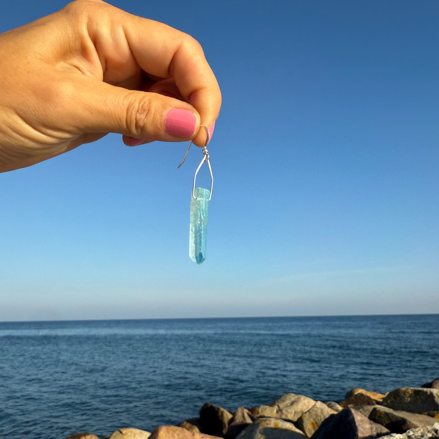 Hand holding a blue crystal earring against a blue sky and ocean background