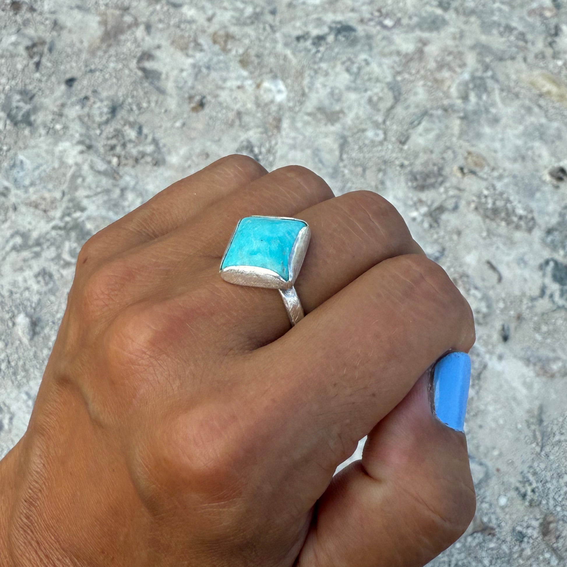 Hand wearing a turquoise square stone ring on a textured surface