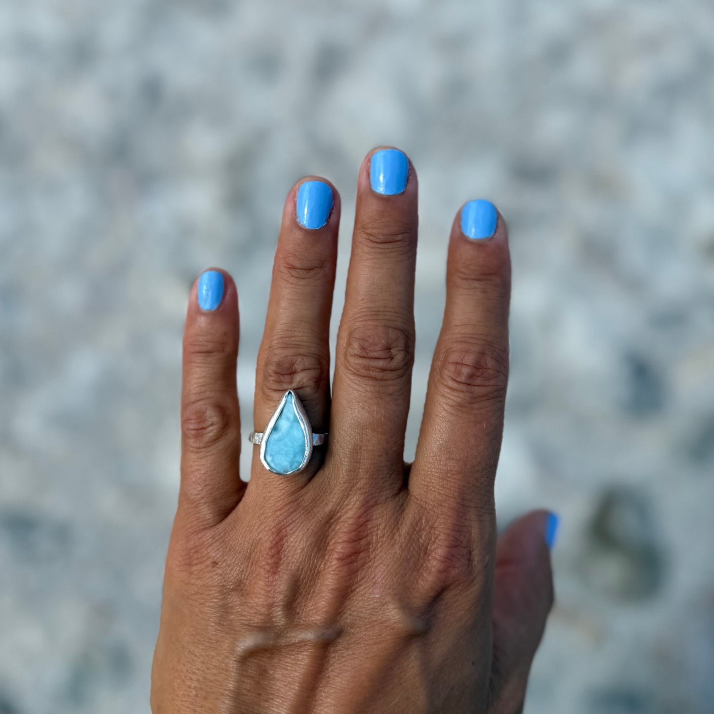Hand with blue nail polish wearing a silver ring with a pearshaped larimar gemstone against a blurred background