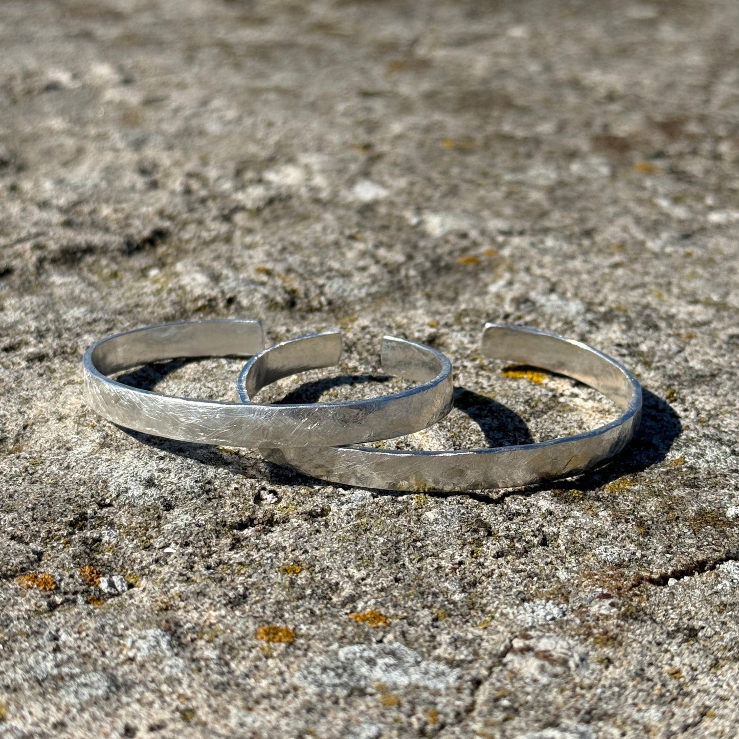 two silver cuffs in brushed sterling silvere on a textured stone surface