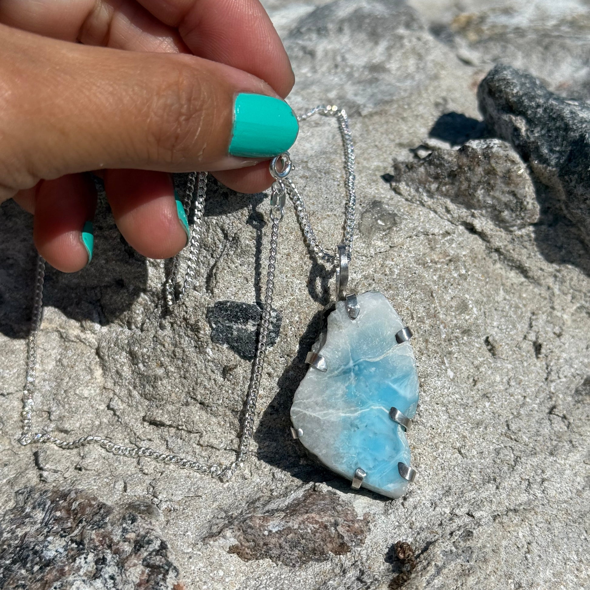 Heart-shaped blue stone pendant held by a hand with turquoise nail polish on a rocky surface.