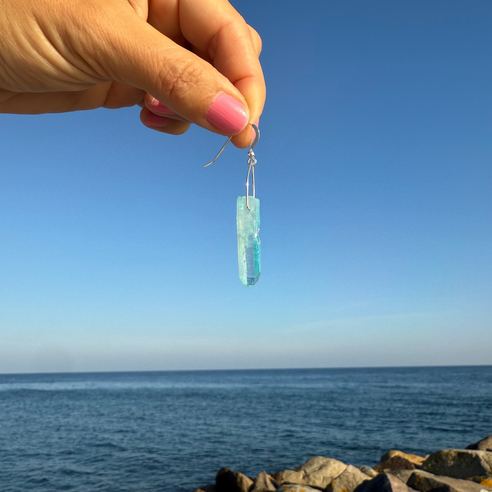 Hand holding a blue teardrop-shaped earring with a clear blue sky and ocean background