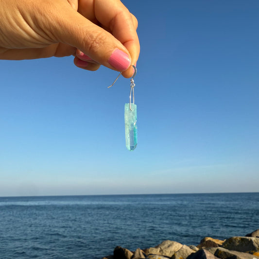 Hand holding a blue teardrop-shaped earring with a clear blue sky and ocean background