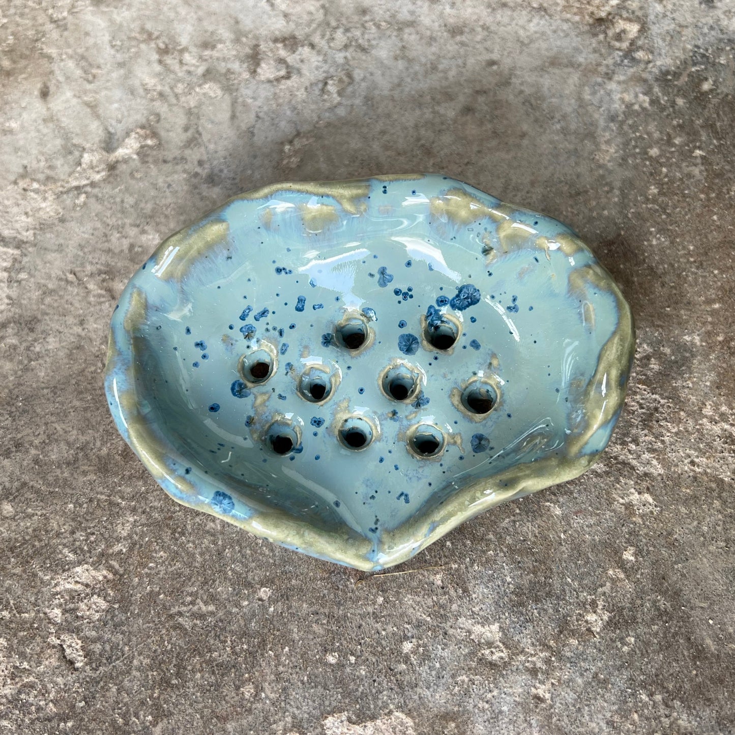 Blue ceramic soap dish in the form of a seashell. The perfect amount of holes to keep the soap bar fresh over time. Seashell soap dish on gray concrete.