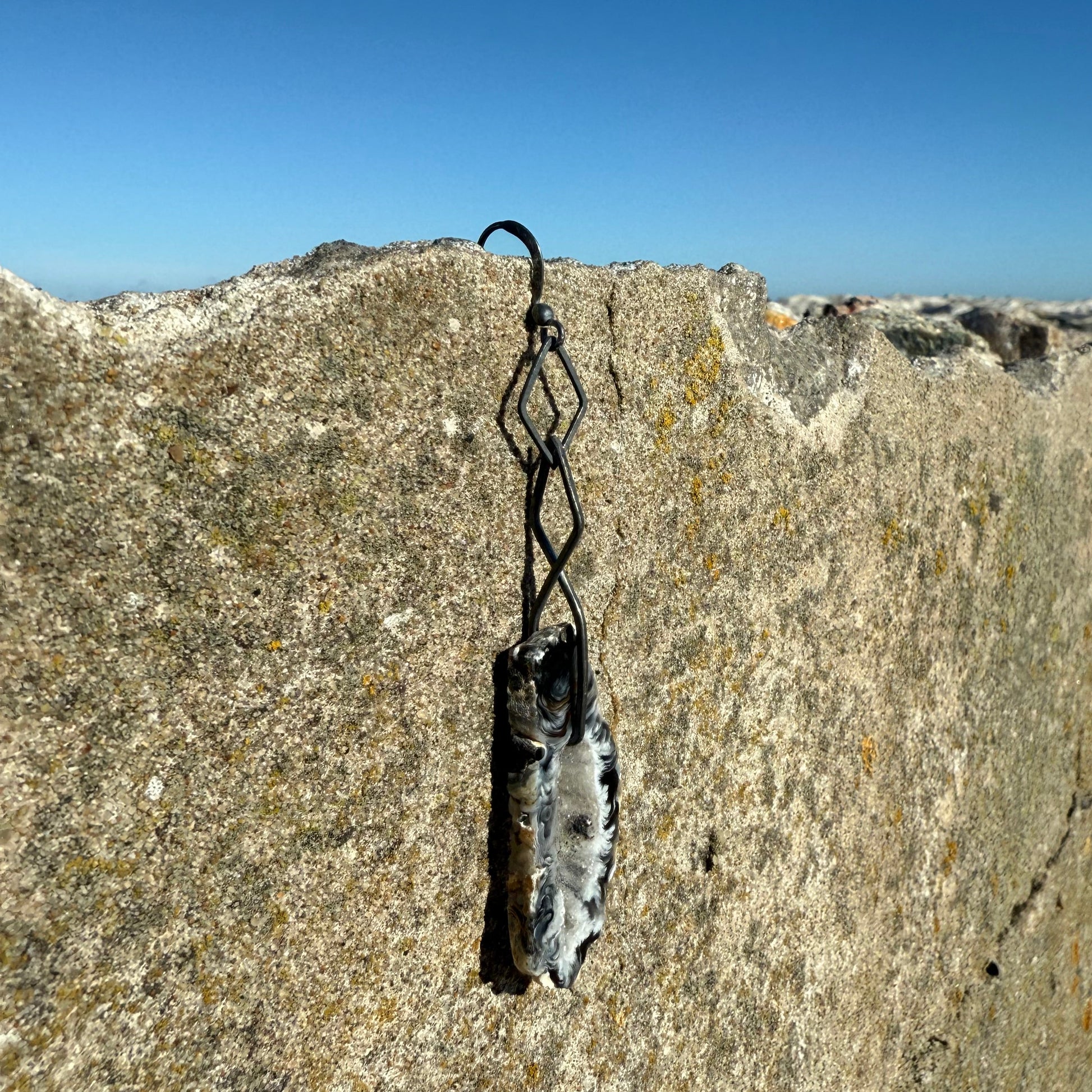 black agate crystal slice earring in oxidized silver hangin on a pier by the sea with blue sky in the background