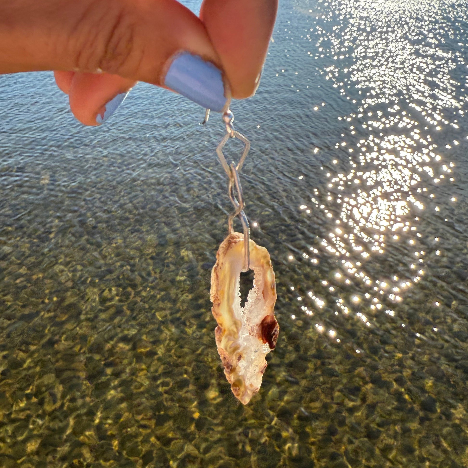 Hand holding a dangling silver arring with raw crystal on a natural background of glittery water and sky.