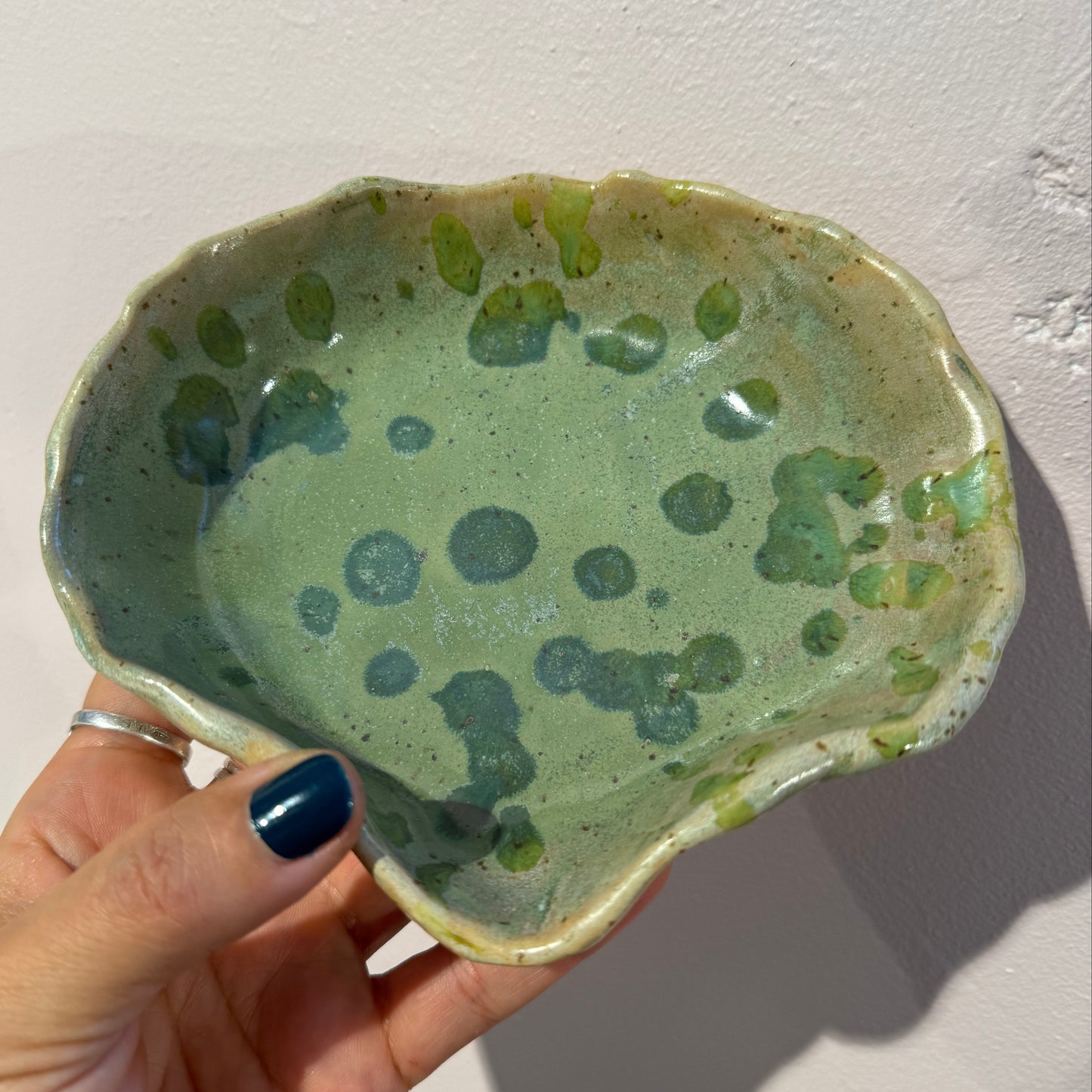 Hand holding a green seashell ceramic dish with speckled pattern against a soft pink background