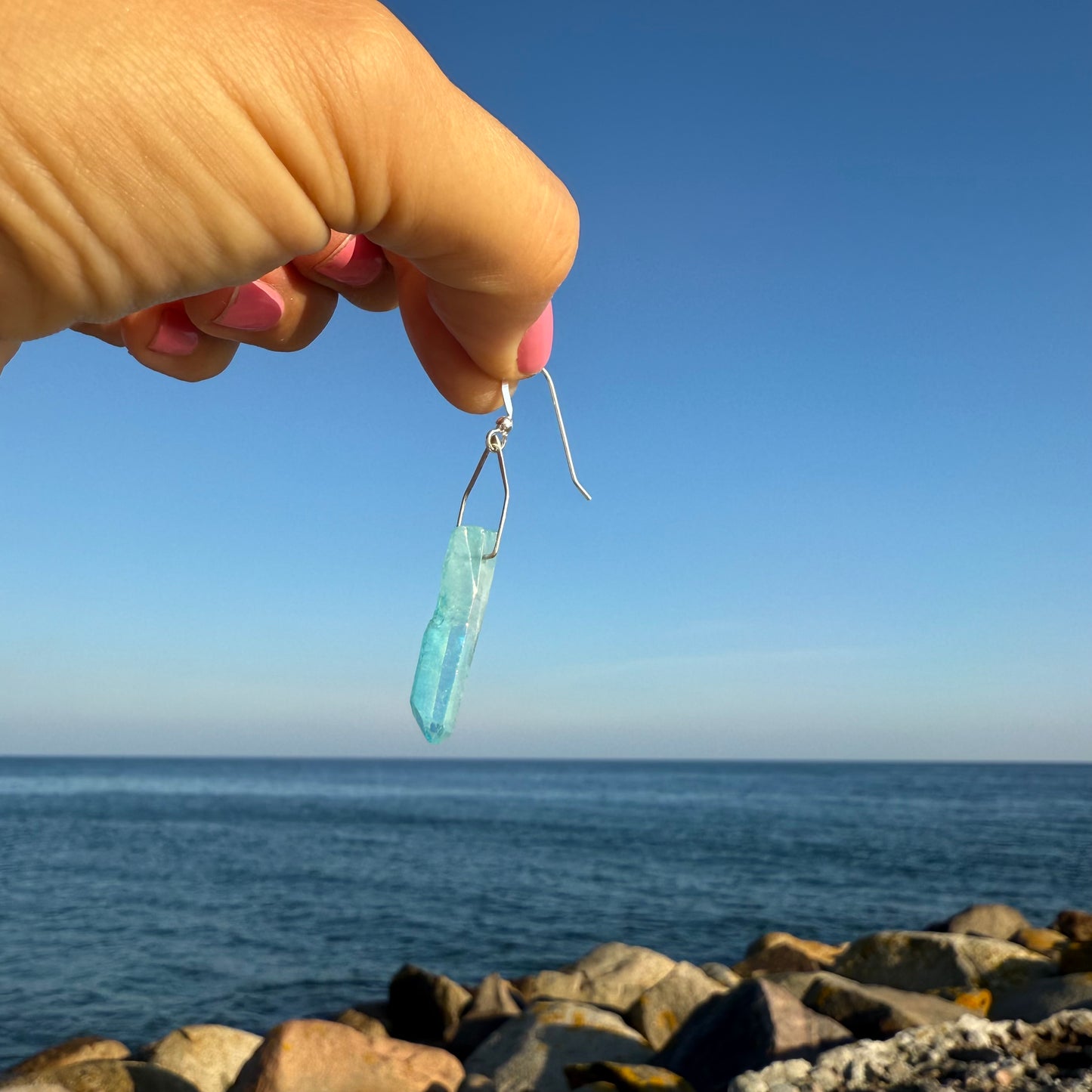 Turquoise teardrop earring held by a hand with a clear blue sky and ocean background