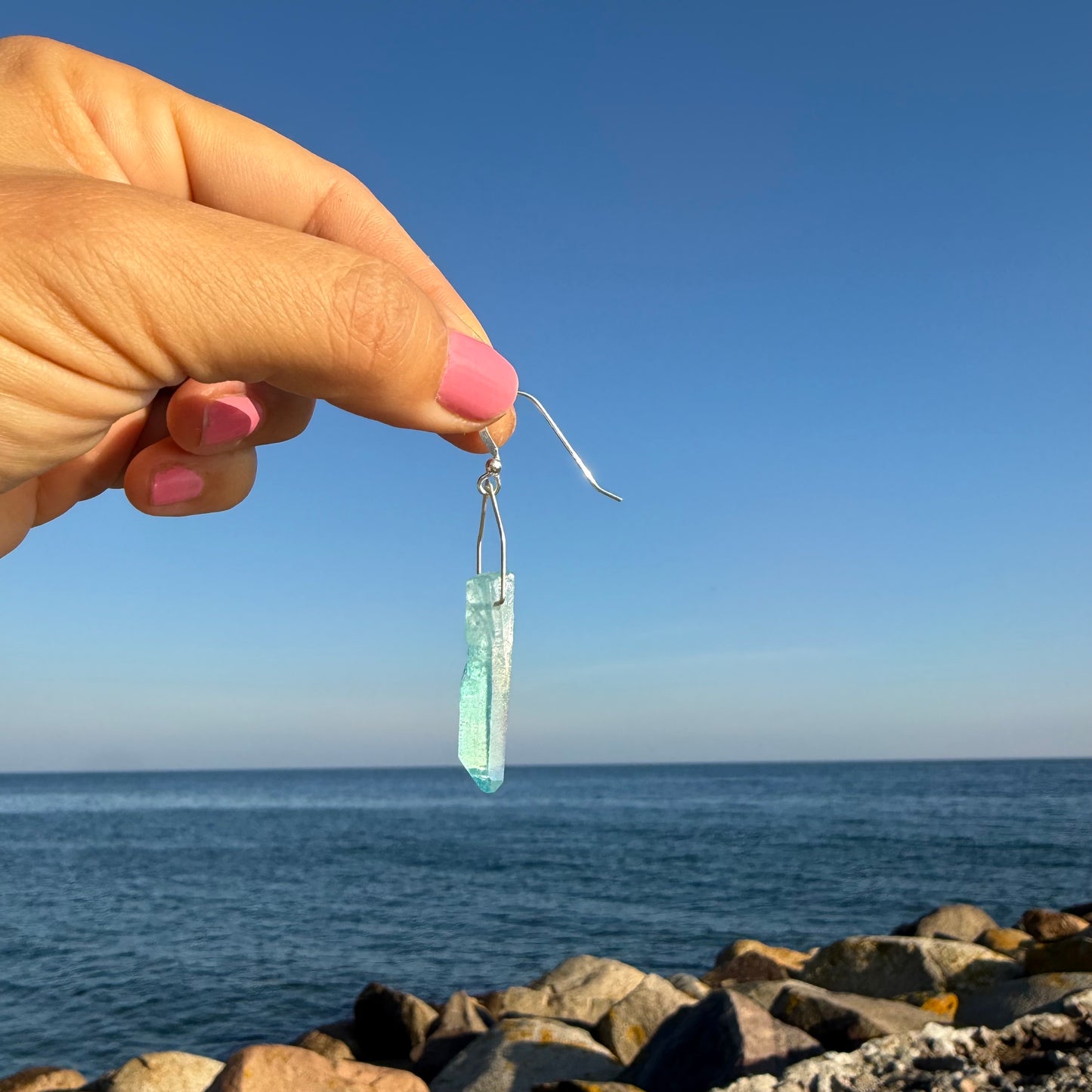 Hand holding a clear aqua blue titanium quartz mono silver earring against a clear blue sky and ocean background