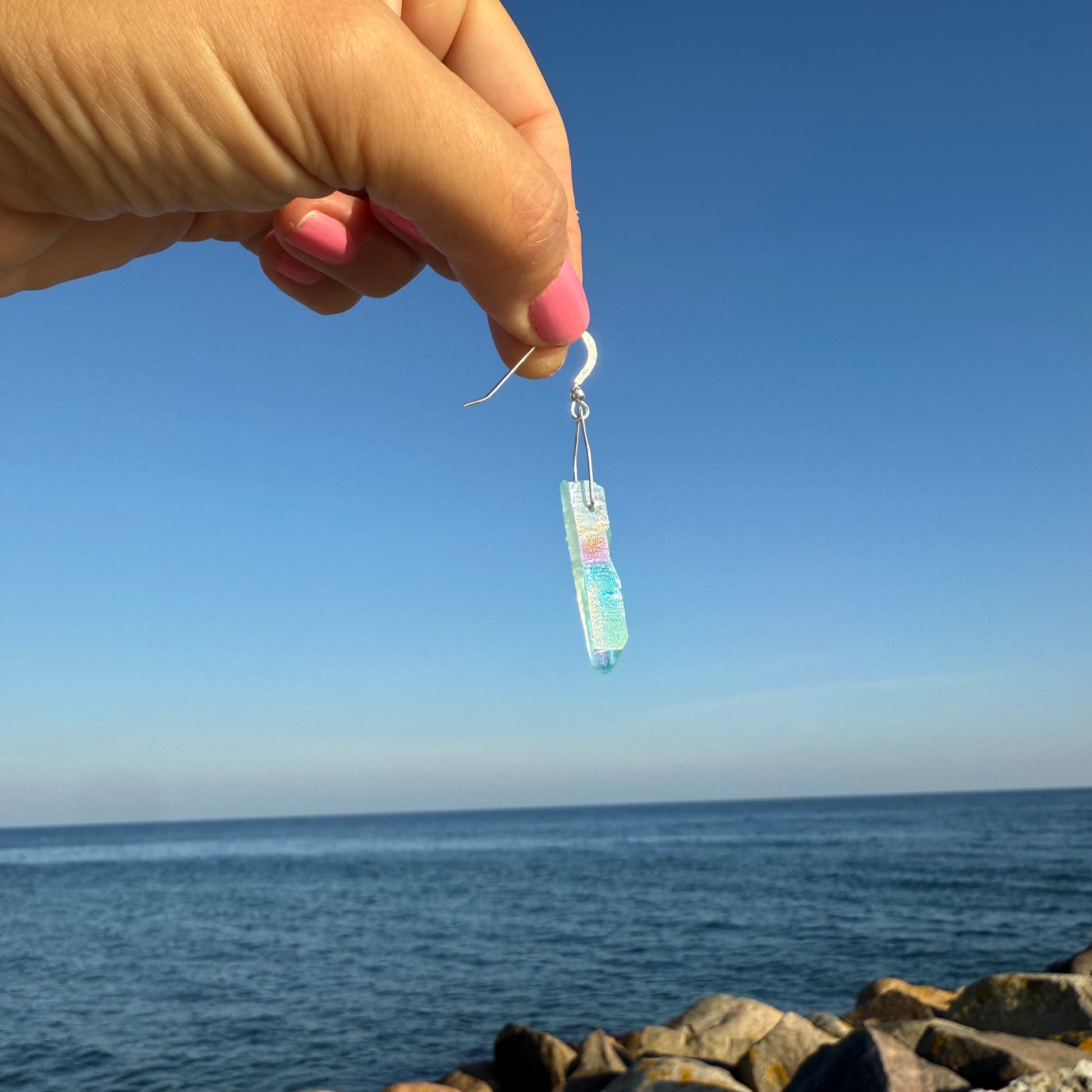 Earring with a colorful dangling crystal earring held by a hand against a blue sky and ocean background