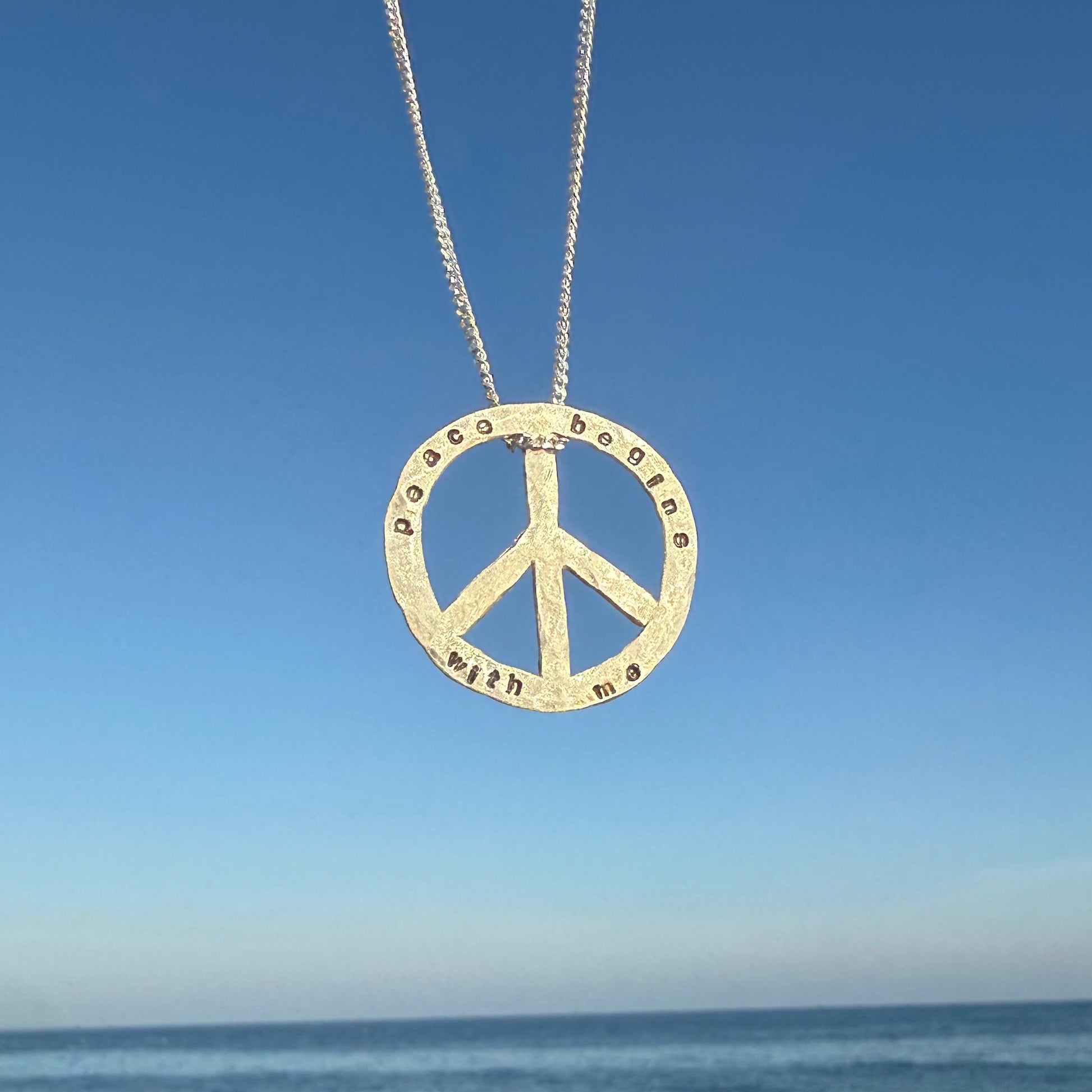 Peace sign necklace with engraved text "peace begins with me" against a blue sky and ocean background