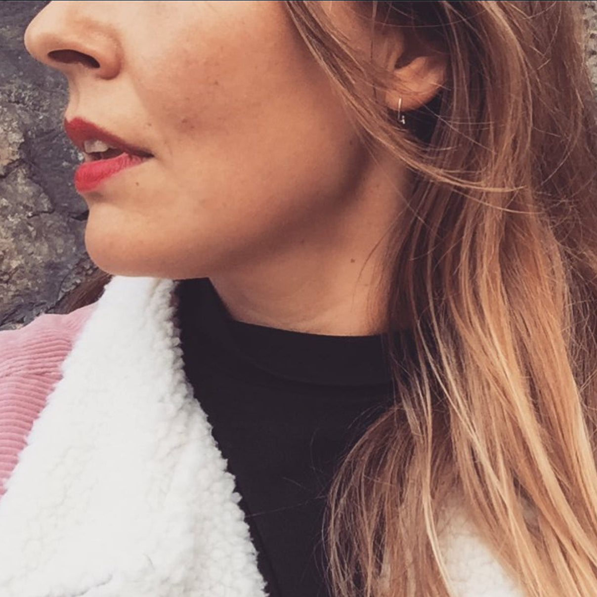 Close-up of a woman with petite silver hoop earring, red lipstick and blonde hair, wearing a black top and white jacket.