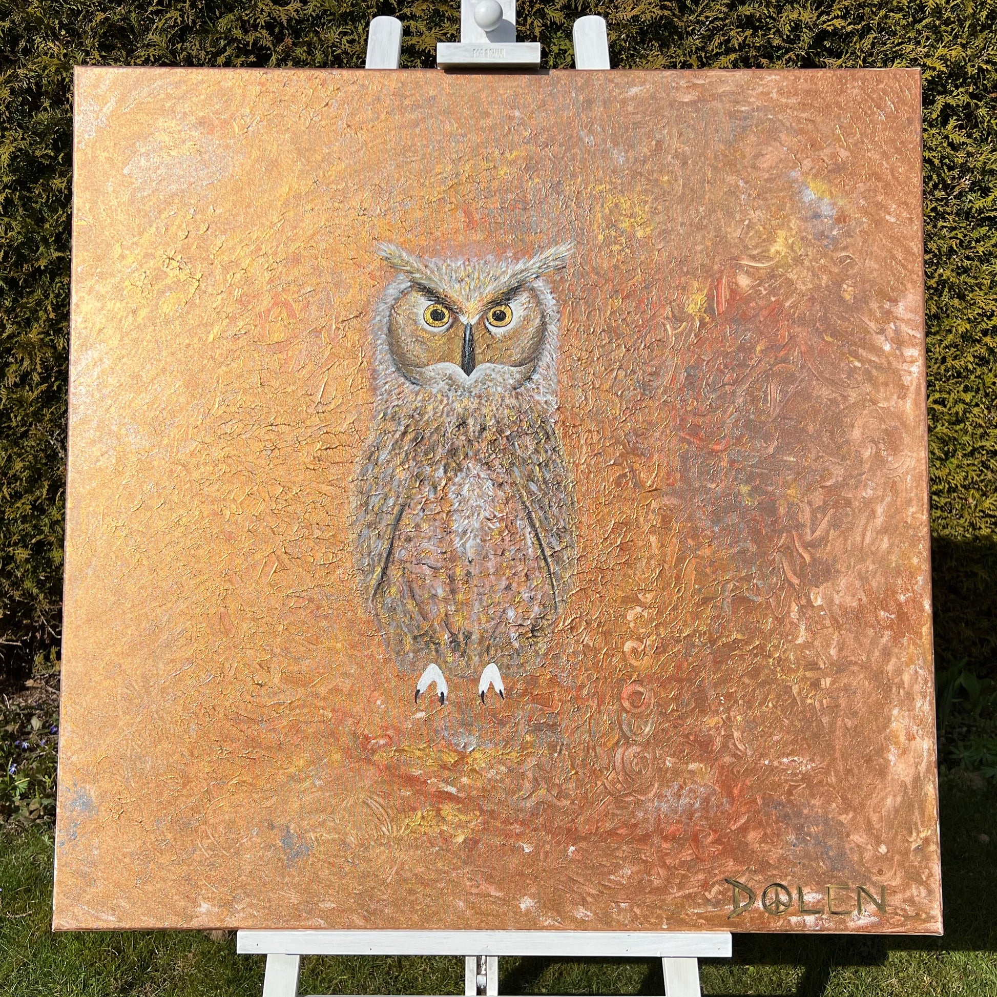 Painting of an owl on a textured gold background, displayed on an easel outdoors.