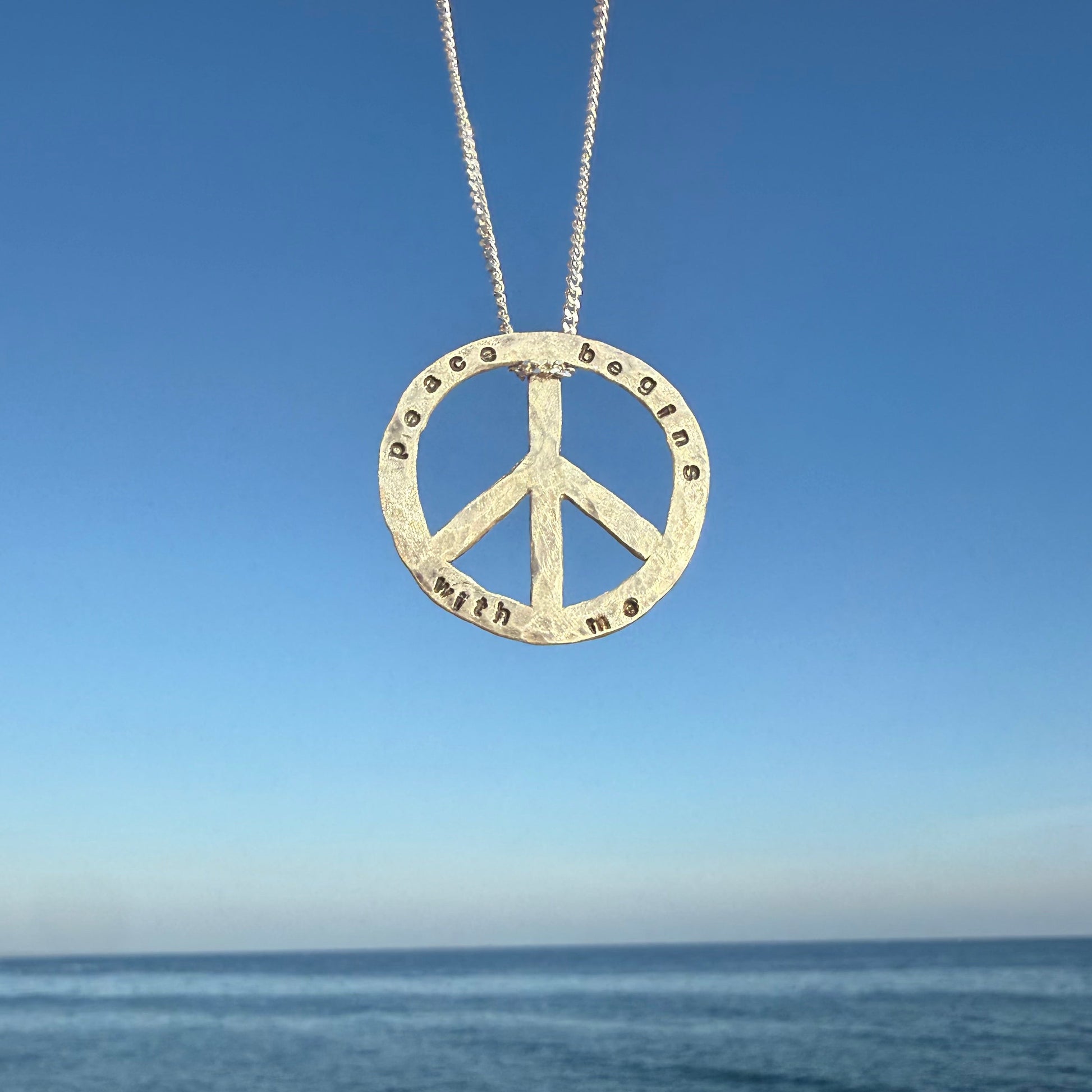 Peace symbol necklace with engraved text against a blue sky and ocean background