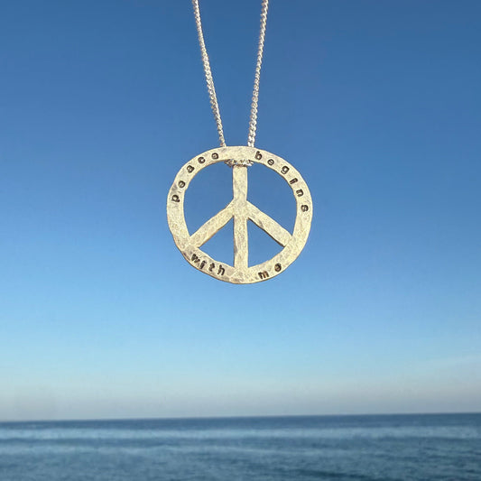 Peace symbol necklace with engraved text against a blue sky and ocean background